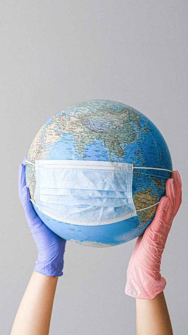 World globe with a mask, held by a gloved hand on one side and a bare hand on the other, symbolizing global health dynamics during COVID-19.